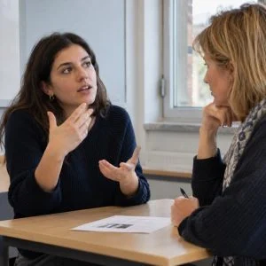 Scena illustrativa generata con AI: una candidata si esercita per lo speaking con una docente.